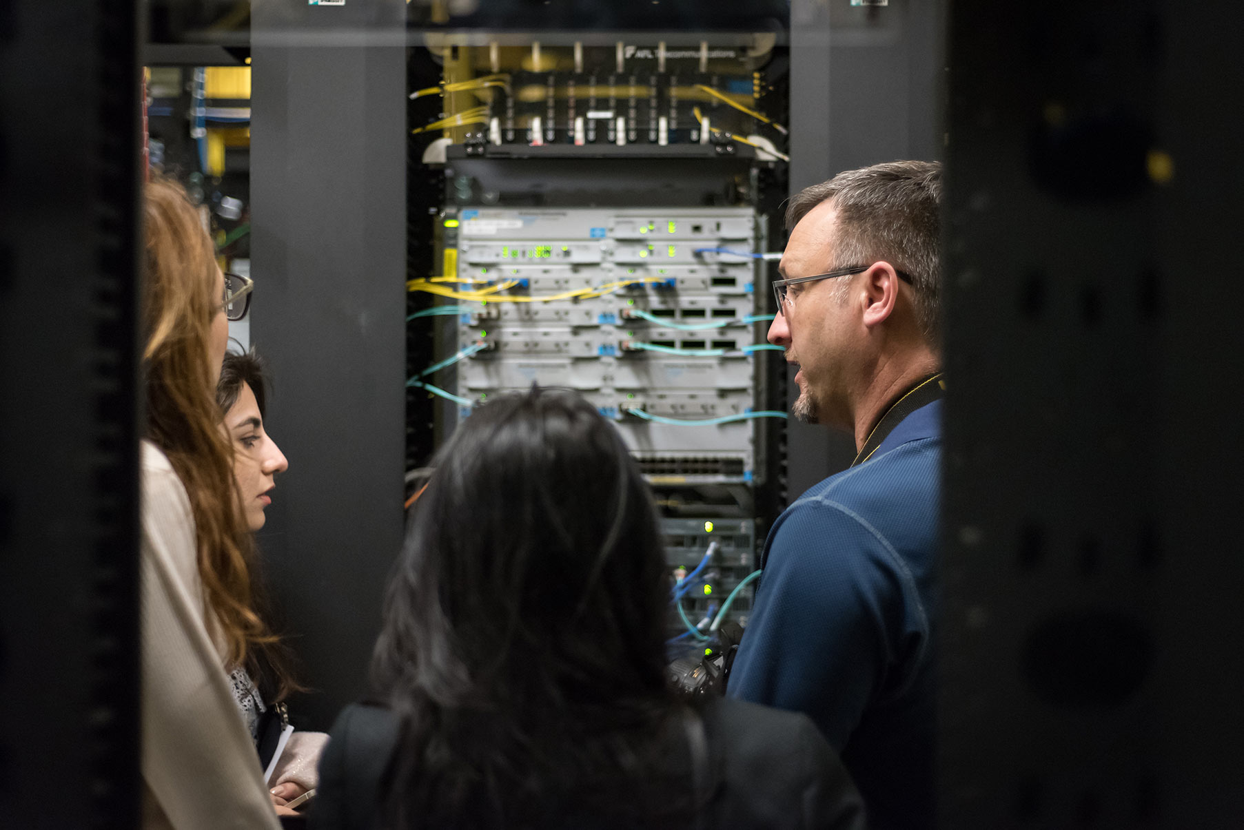 A man gives a data center tour to three women. He is talking and they are all looking at a large server rack.