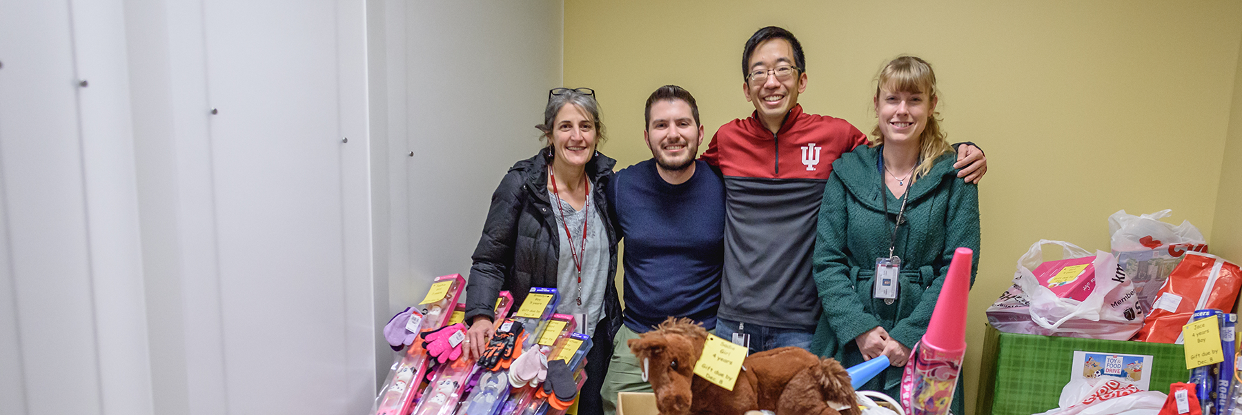 four UITS staff members (two women and two males with arms around each other) stand amidst a bunch of toys they have arranged to deliver to needy children