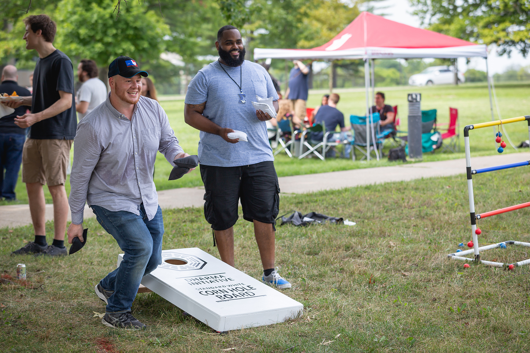 two men standing side by side at a staff picnic, holding corn hole bean bags, one of them beginning to throw his. both are smiling.